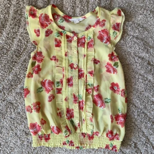 Blommig gul blus från Pumpkin Patch - Söt gul blus från Pumpkin Patch med röda blommor och volangdetaljer. Blusen har knappar framtill och korta ärmar. Perfekt för en solig dag!