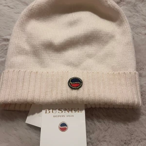 Vit mössa från Busnel - Elegant vit mössa från Busnel med ribbad kant och ett stilrent emblem framtill. Perfekt för att hålla värmen med stil under kyliga dagar. Onesize.