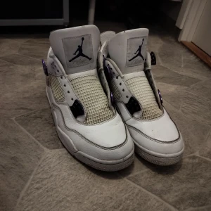 Jordan 4 metallic purple  - Snygga vita Nike Air Jordans med lila detaljer. Skorna har en klassisk design med snörning och perforerade paneler för ventilation. Den ikoniska Jumpman-loggan pryder både tungan och hälen. Perfekta för dig som vill ha en stilren look med en touch av färg.