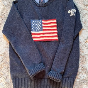 Mörkblå stickad tröja från GANT - Säljer en snygg mörkblå stickad tröja från GANT med ett stort amerikanskt flaggmotiv på framsidan. Tröjan har långa ärmar och ribbade muddar vid ärmslut och nederkant. Perfekt för en snygg look! Jag är 154 cm och tröjan är lite för stor för mig. Skulle säga att den passar M om man vill att den ska sitta lite snyggt. Tveka inte om att höra av er om eventuella funderingar!🤝 Priset kan också diskuteras! 