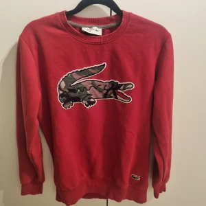 Röd tröja från Lacoste - Röd tröja från Lacoste med en stor kamouflagekrokodil på bröstet. Tröjan har långa ärmar och ribbade muddar vid ärmslut och nederkant.