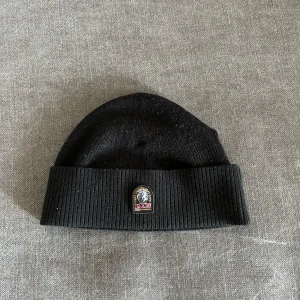 Svart ribbad mössa med broderad logga - Snygg svart ribbad mössa med en cool broderad logga framtill. Perfekt för att hålla värmen och stilen under de kallare månaderna. Enkel att matcha med olika outfits.