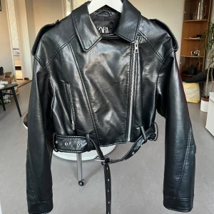 Svart skinnjacka från Zara - Snygg svart skinnjacka från Zara med dragkedja och bälte i midjan. Jackan har en klassisk bikerstil med långa ärmar och axelklaffar. Perfekt för en cool look!