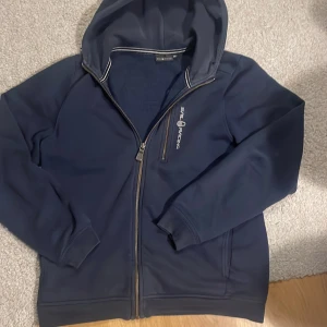 Mörkblå hoodie från Sail Racing - Säljer en snygg mörkblå hoodie från Sail Racing med dragkedja och huva. Perfekt för kyliga dagar. Den har långa ärmar och en stilren design med logotyp på bröstet. Passar både till vardags och mer avslappnade tillfällen. Tyvärr finns det ett litet hål vid luvan som man inte tänker på. Pris går att diskuteras.