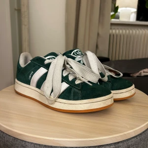 Gröna Adidas Campus 00s sneakers - Snygga gröna Adidas Campus sneakers med vita detaljer och breda snören i storlek 39. Köptes efter sommaren och har inte kommit till användning nu under hösten/vintern så inte mycket alls använda. Super fint skick, inte något slitna/sönder alls.