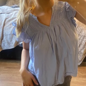 Ljusblå blus med volangdetaljer - Charmig ljusblå klänning med volangdetaljer på ärmarna och v-ringad hals. Perfekt för en somrig look med sin luftiga design och mjuka material. Står strl 158 / 164 i den, men skulle säga att den är som en xxs / xs