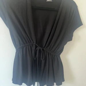 Svart omlottopp från Ginatricot - Snygg svart omlottopp från Ginatricot med korta ärmar och knytband i midjan. Perfekt för en stilren look. Aldrig använd 