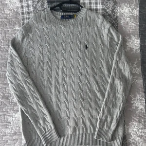 Grå stickad tröja från Polo Ralph Lauren - Säljer en stilren grå stickad tröja från Polo Ralph Lauren med det klassiska broderade logotypen på bröstet. Tröjan har ett snyggt kabelstickat mönster och långa ärmar. Perfekt för kyligare dagar!