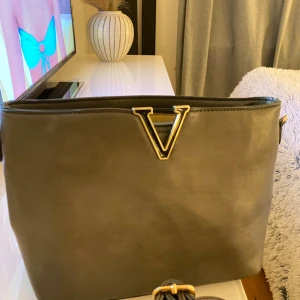 Grå väska med gulddetaljer - Snygg grå väska med ett stort V i guld på framsidan. Man får med sig ett band till med. Vet ej märket för köpte på busfröt (en secondhandbutik). Skriv vid frågor eller intresse!