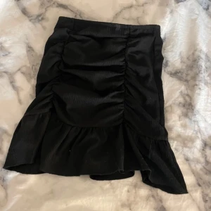 Svart kjol med volangdetaljer - Snygg svart kjol med volangdetaljer och en smickrande passform. Kjolen har en kort längd och är perfekt för att ge en elegant touch till din outfit. Den är tillverkad i ett mjukt material som ger en bekväm känsla.