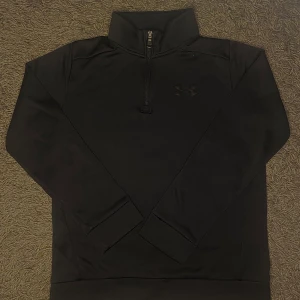 Svart tröja med dragkedja från Under Armour - Säljer en svart tröja från Under Armour med halv dragkedja och hög krage. Tröjan har en lös passform och är perfekt för kyligare dagar. Den är tillverkad i ett mjukt material och har en diskret logga på bröstet. Denna tröjan har inga skador