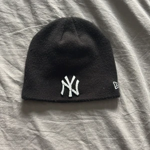 Svart mössa från New Era - Snygg svart mössa från New Era med broderad logga framtill. Perfekt för att hålla värmen under kyliga dagar. Den har en klassisk design som passar till det mesta.