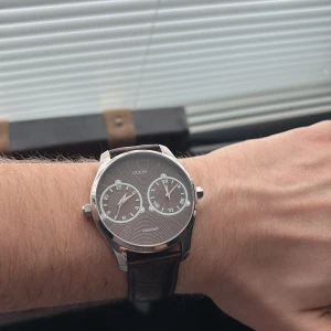 Stilren klocka från Guess - Snygg klocka från Guess med en silverfärgad boett och en brun skinnrem. Urtavlan är grå med romerska siffror och två mindre urtavlor. Perfekt för den som vill ha en elegant accessoar.