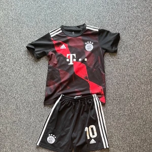Svart och röd fotbollströja med shorts bayern - Snygg fotbollströja från Adidas i svart och rött med korta ärmar. Tröjan har ett coolt geometriskt mönster och klubbmärke på bröstet. Matchande svarta shorts med vita ränder och nummer 10. Perfekt för fotbollsälskare! Lite sliten