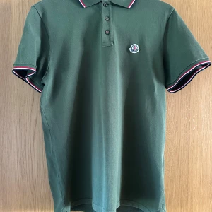 Moncler piké - Snygg grön pikétröja från Moncler med korta ärmar och klassisk krage. Tröjan har en broderad logotyp på bröstet och röd-vita detaljer på krage och ärmslut. Perfekt för en stilren look. Kvitto finns!