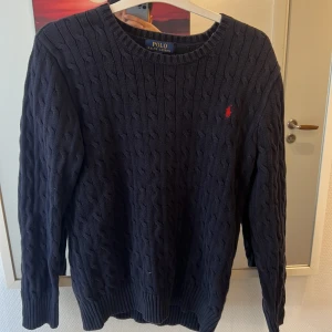 Mörkblå stickad tröja från Ralph Lauren - Säljer en snygg mörkblå stickad tröja från Ralph Lauren med kabelstickat mönster. Tröjan har långa ärmar och en liten röd logotyp på bröstet. Perfekt för kyligare dagar!