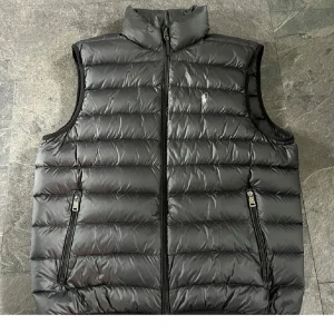 Svart dunväst från Polo Ralph Lauren - Svart dunväst från Polo Ralph Lauren med klassisk broderad logga på bröstet. Västen har horisontella quiltade paneler, hög krage och två praktiska fickor med dragkedja. Perfekt att slänga över en hoodie för extra värme och stil.