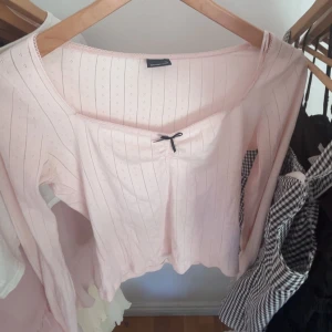 Ljusrosa topp med rosett från Gina Tricot - Supersöt ljusrosa topp från Gina Tricot i storlek M. Den har lång ärm, v-ringad hals med liten svart rosett och diskret hålmönster med tunna ränder. Materialet är mjuk bomull och toppen har en feminin, normal passform. Perfekt för dig som gillar gulliga detaljer!