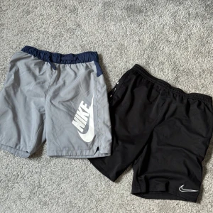 Nike shorts 2-pack - Två par Nike shorts, ett par svarta med små vita swoosh längs midjan och ett par gråa med marinblå midja och stor vit Nike-logga på benet. Storlek 164. Båda har elastisk midja och är gjorda i lätt polyester, perfekta för träning eller chill.