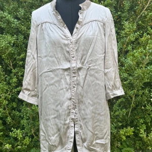 Beige lång skjorta med spetsdetaljer - Snygg beige skjorta i silkesliknande material med trekvartsärm och dekorativa knappar framtill. Spetsdetaljer längs knappraden och axlarna ger en cool vintagekänsla. Perfekt för dig som gillar en lite längre och loose passform. Knappar fattas.