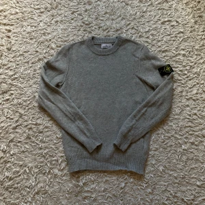Grå stickad Stone Island tröja - Säljer en grå stickad tröja i ull från Stone Island med klassisk rund hals och ribbade muddar. Tröjan har lång ärm och den ikoniska Stone Island patchen på vänster arm. Perfekt för dig som gillar clean och stilren streetwear. Den är helt ny och oanvänd, originella tags medföljer vid köp. Passar dig som är 175-185 (passar lite baggy för storlek)