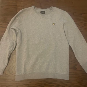 Grå sweatshirt från Lyle & Scott - Snygg grå sweatshirt från Lyle & Scott Junior med klassisk rund hals och långärmad design. Tröjan har en liten gul broderad logga på bröstet och är tillverkad i mjuk bomullsmix som är skön att ha på sig. Perfekt för chill dagar och enkel att matcha med jeans eller joggers.