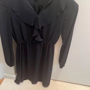 Svart klänning med volang och lång ärm - Det Snygg svart klänning med lång ärm och volangdetalj vid halsen. Klänningen har en markerad midja med resår och ett mjukt, flowy tyg som ger en skön passform. Perfekt för dig som gillar stilrena plagg med en twist.