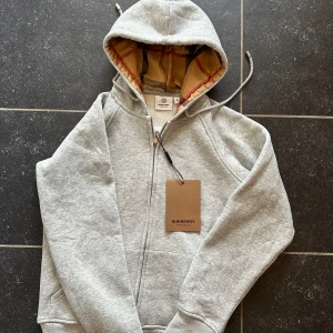 Grå Burberry hoodie med rutig huva - Säljer en grå hoodie från Burberry, passar som en XS ksk mindre skulle säga för en max 13åring, Tröjan har dragkedja, snörning och en ikonisk rutig huva i beige, rött och svart. Mjuk bomullskänsla och klassisk Burberry-vibe. Perfekt för dig som vill ha en clean look med lyxig touch.