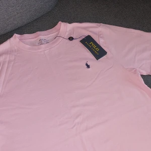 Ljusrosa t-shirt från Polo Ralph Lauren - Säljer en ljusrosa t-shirt från Polo Ralph Lauren med klassisk broderad logga på bröstet. T-shirten har rund halsringning och är tillverkad i mjuk bomull. Perfekt för en avslappnad och stilren look.