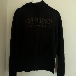 Svart hoodie från Hugo Boss - Svart hoodie från Hugo Boss med guldprickigt BOSS-tryck på bröstet. Tröjan har huva med snörning, stor magficka och långa ärmar med ribbade muddar. Perfekt för dig som gillar stilrena och lyxiga streetwear-plagg.