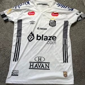 Santos FC Neymar Jr. 10 matchtröja - Snygg vit Santos FC fotbollströja från Umbro med Neymar Jr. och nummer 10 på ryggen. Tröjan har korta ärmar, svarta tryck och flera sponsorer som blaze.com och HAVAN. Materialet är lätt och andas, perfekt för träning eller match. Coola svarta detaljer längs sidorna och ärmarna.