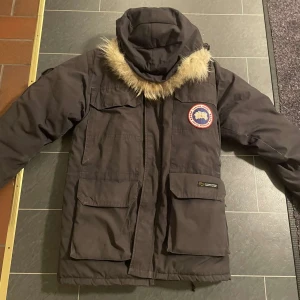 Canada Goose mörk parkajacka med päls - Säljer en mörkbrun Canada Goose parkajacka med ikonisk patch på ärmen och fluffig päls runt huvan. Jackan har stora fickor framtill, dragkedja och knappar, perfekt för kalla dagar. Snygg och funktionell design som sticker ut.