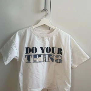 Vit t-shirt med tryck från Lindex - Vit t-shirt från Lindex med coolt tryck 'DO YOUR THING' på framsidan. Bokstäverna har svart och blått camouflagemönster. Modellen är kortärmad och har en klassisk rund hals. Materialet är mjuk bomull, perfekt för chill dagar.