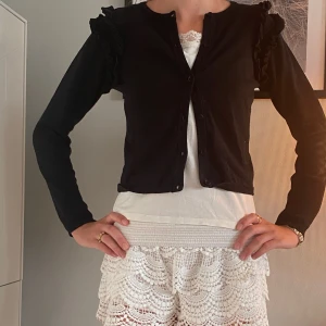 Svart kofta med volangdetaljer - Svart kofta med rund hals och knappar framtill. Långärmad modell med snygga volangdetaljer på axlarna som ger en cool och trendig vibe. Perfekt att slänga över en topp för en chill look. Materialet känns mjukt och av bomull.