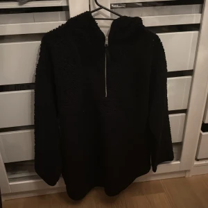 Svart fleece hoodie med half zip - Mysig svart hoodie i fleece med huva och half zip framtill. Tröjan har långärmade ärmar och en fluffig, teddy-liknande yta som gör den extra skön och varm. Perfekt för kalla dagar när du vill ha något riktigt bekvämt och chill. Passar även S om du vill ha den oversized.