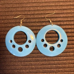Blå vintage  plastörhängen - Säljer ett par coola blå örhängen i rund form med flera cirkulära hål som ger en retro vibe. Perfekt för dig som vill sticka ut med ett unikt och färgglatt smycke. Passar till alla som gillar statement-accessoarer.