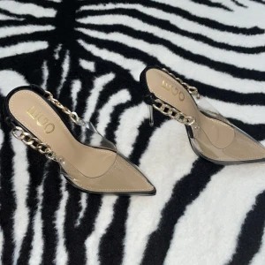 Transparenta pumps med guldkedja från EGO - Säljer ett par riktigt snygga pumps från EGO med transparent plast och spetsig tå. Skorna har en cool guldfärgad kedja som detalj vid hälen och en hög, klar klack. Svarta kanter och beige innersula ger en stilren vibe. Perfekt för dig som vill sticka ut med din look.