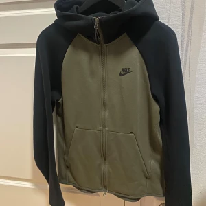 Nike Tech Fleece - Nike hoodiejacka i svart och olivgrön med dragkedja framtill och två stora fickor. Jackan har huva och långärmade svarta ärmar, samt Nike-logga på bröstet. Perfekt för chill eller träning, tillverkad i mjuk bomull/polyesterblandning.