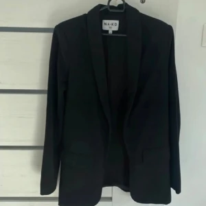 Svart kavaj från NA-KD - Stilren svart kavaj från NA-KD med klassisk krage och två fickor framtill. Modellen är rak och har långa ärmar, perfekt för dig som gillar en clean och enkel look. Materialet är mjukt och känns bekvämt mot huden.