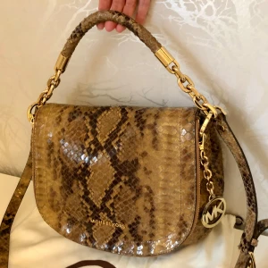 Michael Kors väska med ormskinnsmönster - Michael Kors-väska i glansigt brunt ormskinnsinspirerat mönster med guldfärgade detaljer och diskret MK-logga. Väskan är i mycket gott skick, knappt använd. Skick: Nästan ny, använd några gånger Bäralternativ: Kan bäras över axeln; två avtagbara handtag (kort och långt) som går att kombinera efter behov Design: Elegant och tidlös modell som fungerar både till vardag och fest