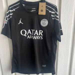 PSG svart matchtröja Dembele #10 - Snygg svart PSG fotbollströja med vita detaljer, Jordan-logga och Qatar Airways tryck på bröstet. Axelpartiet har coola vita linjer och på ryggen står O. Dembélé med nummer 10. Materialet är lätt och andas, perfekt för match eller träning.