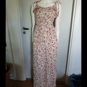 Blommig långklänning med knytband - Superfin långklänning med blommigt mönster i rött och grönt på vit botten. Klänningen har smock i midjan, volangdetaljer vid axlarna och knytband på axlarna. Perfekt för sommarens alla tillfällen, luftig och romantisk stil.