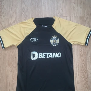 Sporting CP CR7 fotbollströja Nike S - Sporting CP fotbollströja med CR7-tryck från Nike i Dri-Fit material. Svart kropp med guldiga korta ärmar och detaljer, klubbmärke broderat på bröstet och Betano sponsortryck. Perfekt för dig som gillar snygg design och fotboll.