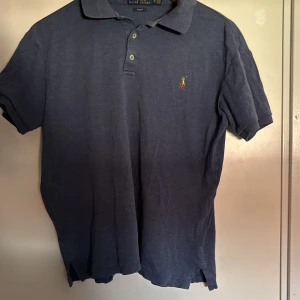 Blå Polo Ralph Lauren t-shirt slim fit till barn  - Snygg marinblå t-shirt från Polo Ralph Lauren i slim fit-modell. Klassisk pikékrage, korta ärmar och två knappar framtill. Ikonisk broderad logga på bröstet. Tillverkad i mjuk bomull som är skön mot huden. Till barn 