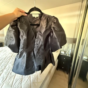 Svart blus med puffärm By Malina XS - Supersnygg svart blus från By Malina i storlek XS. Blusen har puffiga korta ärmar, v-ringad hals och dekorativa knappar framtill. Materialet är glansigt och känns lyxigt, perfekt för dig som gillar statement-plagg med cool siluett.