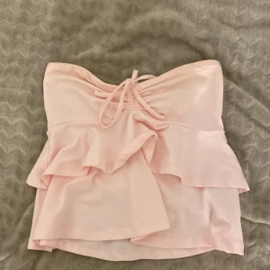 Ljusrosa bandeau topp från Gina Tricot - Supersöt ljusrosa bandeau topp från Gina Tricot med volangdetaljer och snörning framtill. Toppen är axelbandslös och har en mjuk, flowy form som ger en romantisk vibe. Perfekt för dig som vill ha en feminin och trendig look.