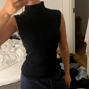 Svart polotopp ärmlös - Snygg svart polotopp med hög krage och ärmlös design. Toppen har en slim passform som sitter tajt mot kroppen och ger en clean look. Perfekt att styla med jeans eller kjol för en cool vibe.