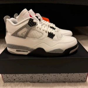 Nike Air Jordan 4 White Cement sneakers - Nike Air Jordan 4 White Cement sneakers med vit ovandel i skinn, grå och svart detaljer samt röd logga på plösen. Sulan har grått cementmönster och röda Nike-detaljer undertill. Klassisk meshpanel på sidorna och snörning framtill. Perfekt för dig som gillar retrostil och streetwear.