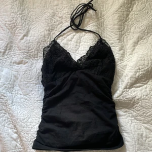 Svart spetsig halterneck-topp från SHEIN - Svart halterneck-topp från SHEIN i storlek XS. Toppen har v-ringad spets upptill och tunna knytband i nacken. Materialet är mjukt och stretchigt, med en slim passform som sitter snyggt mot kroppen. Perfekt för dig som gillar en feminin och trendig look.
