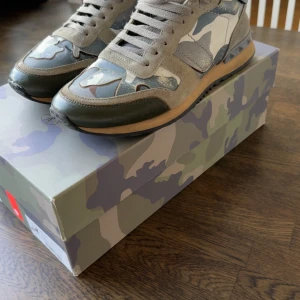 Valentino Rockrunner  - Snygga Valentino Rockrunner sneakers med grått, grönt och beige camouflage-mönster. Skorna har detaljer i mocka och skinn, rund tå, snörning och platt sula. Baksidan har coola pyramidnitar för extra edge. Perfekt för dig som gillar streetstyle och designer.